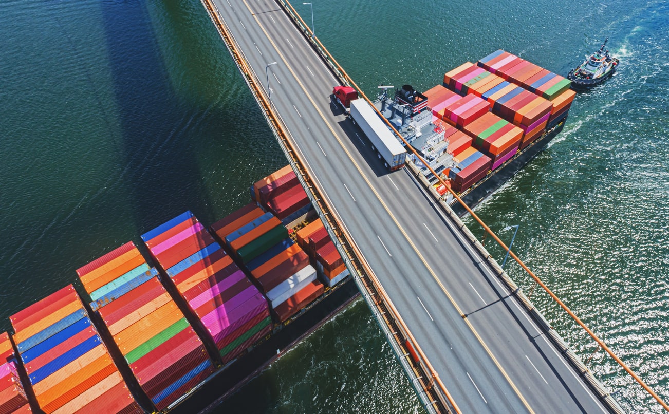 Aerial drone view of a container ship navigating beneath a semi truck crossing a suspension bridge